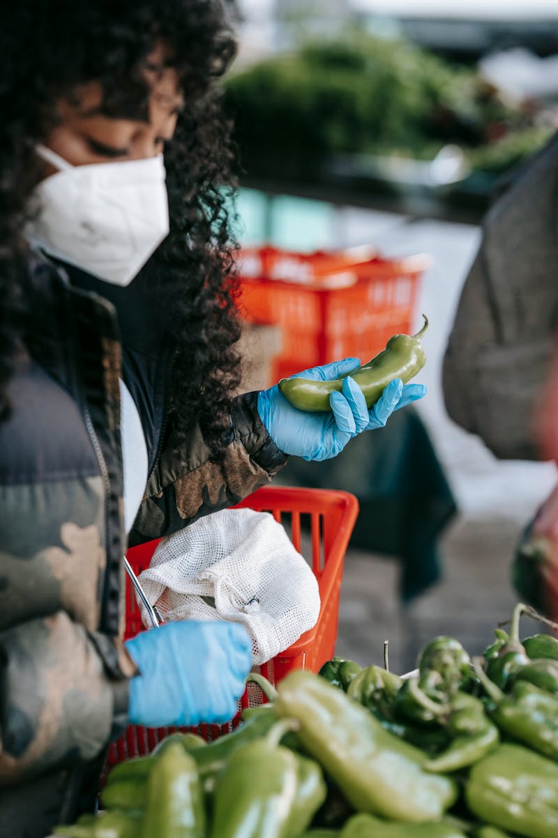 Crop concentrated African American female in respirator and gloves picking ripe capsicums while doing groceries in supermarket