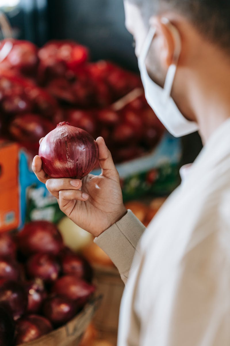 Side view of crop unrecognizable male in casual clothes and medical mask choosing fresh raw red onions in local food market